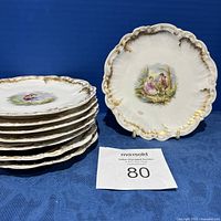 Six Limoges plates stacked showing scalloped edges with gold hand embellishments and a plate displayed upright showing a romantic scene with a couple near foliage.