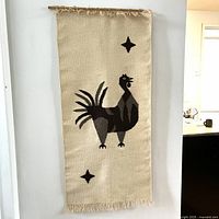 Wool ethnic rug hung on wall showing rooster design with two star shapes on beige background, fringed edges.