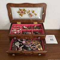 Open wooden jewellery box showing top compartments with sorted earrings, rings, and brooches, with the lower drawer containing mixed bracelets and watches