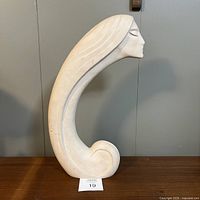 Full side view of large stylized elongated head sculpture with curled hair base, white with scuffs, standing on table.