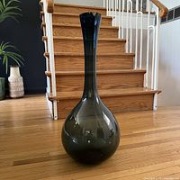 Front view of blue glass floor vase with rounded base and tapered neck standing on wooden floor in front of stairs.