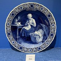 Front view of the large Royal Delft ceramic plate bowl showing the detailed hand-painted scene of a woman knitting beside a basket with a sleeping child, surrounded by a blue floral and scroll border.
