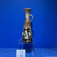 Front angled view of the tall brown speckled vase with abstract painted design and handle against a blue background.