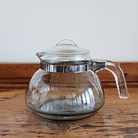 Image showing side view of clear glass Pyrex Flameware teapot with clear glass handle, lid with metal band, on wooden surface.