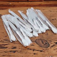 Lot of 12 Noritake Serenade butter spreaders sealed in individual clear plastic sleeves on wooden surface.