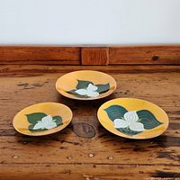 Three graduated enamel on copper bowls with orange-yellow backgrounds and white trillium design arranged on rustic wooden surface