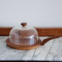 Cheese dome on teak base with handle, clear plastic dome with wooden knob showing glue residue, photographed on marble surface.