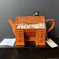 Front view of orange-brown speckled teapot shaped as a roll-top desk with sculpted papers and a tag attached