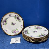 Set of eight 8-inch plates with purple violet floral design and gold rim, stacked and displayed against blue background.