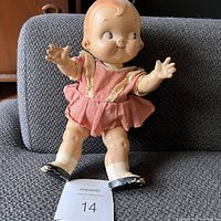 Full front view of vintage composite Campbell's Soup kid girl doll sitting on a grey fabric surface, showing dress and face details with worn paint and patina.