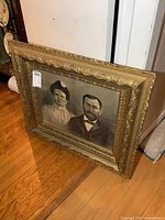 View of the full ornate gold-colored framed portrait leaning against a door, showing the entire artwork.