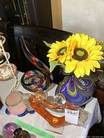 Photo showing heavy glass vase with swirling pattern holding faux sunflowers, glass angel figurine, glass swirl sculpture, and small glass planter on table