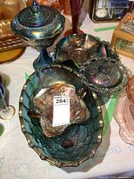 Set of blue carnival glass pieces including two oval footed fruit bowls, a covered candy dish, a serving dish, and a sugar dish with spoon holder, showing iridescent blue finish with floral and grape patterns.