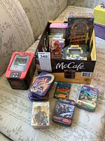 Photo showing a box filled with various collector card tins and packs including American Bandstand box, red Pokemon tin, and others on a couch