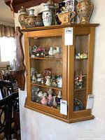 Wall hung wood display cabinet full of small porcelain and ceramic collectibles with large ceramic vases on top