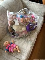 Clear bag filled with multiple small dolls and stuffed toys placed on couch, including a small Cabbage Patch doll lying beside the bag.