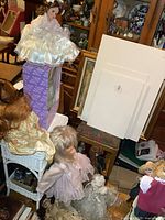 Photo of wedding dress doll sitting on a purple box with several other dolls visible below and around her in a cluttered interior space.