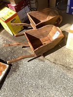 Pair of wooden garden planter wheelbarrows with handles and single wheels positioned on gravel floor next to boxes.
