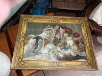 Full view of the framed print with several kittens and floral still life inside an ornate gold frame placed on wooden surface.