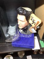 Royal Doulton Elvis Presley character jug bust with attached decorative guitar and teddy bear figurines on a shelf with blue base.