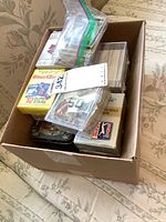 Box filled with various sports cards in plastic bags, cases, and tins showing some card visibility and unopened packs