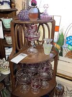 Wooden corner shelf holding two tiers of 4 pink/red stemware glasses with leaf and bird etching, and various perfume bottles on top shelf including Avon Rare Pearls and other vintage bottles.