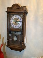 Front view of vintage wooden wall clock hung on wall showing clock face, day-date display, and lower glass pendulum compartment with carved top crest and turned wood columns.