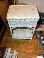 Front view of white vintage wicker stand with lift lid and lower shelf, showing overall shape and woven wicker construction.