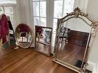 Shows all three mirrors in a room, lined up on the floor by a window. Large fleur de lis topped mirror on the right, oval mirror on the left, and rectangular mirror in the center.