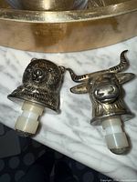Close-up photo of two animal head shaped wine stoppers, one bear and one bull, made of metal with white stopper bottoms, placed on a marble surface and part of a brass item above.