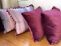 Set of 4 pillows showing 2 maroon velvet and 2 striped silk pillows with pom pom trim on wooden floor.