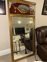 Full front view of large rectangular mirror showing ornate gold frame and wood panel with painting at top.