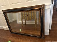 Full view of large rectangular mirror leaning against wall, showing the entire ornate frame and reflective glass surface.