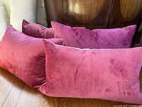 Four rectangular pillows with pink velvet fronts and tan woven cotton backs, arranged on a wooden floor and leaning against furniture.