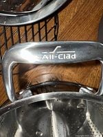 Close-up of All-Clad handle on a roasting dish or steamer insert, showing branded metal handle and part of perforated steamed insert.