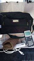 Photo showing black overnight bag, TI-83 Plus calculator, and brown heating pad with controller in front of the bag.