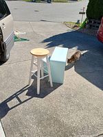 Two waterfall style tables nested and a white legged stool with natural wood seat on concrete driveway under sunlight.
