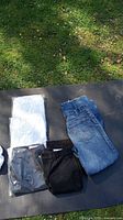 Photo showing two pairs of black pants (one pair in packaging), one pair of Levi's blue denim jeans, and two pinstripe shirts in sealed packaging all laid out on a surface outdoors.