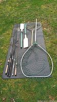 Fishing lot laid out on the grass showing four fishing rods, two oars with green handles, and two fishing nets, one large and one medium size.
