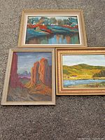 Three framed landscape paintings laid on textured ground: a river with fall foliage, red rock desert, and hilly countryside with river.