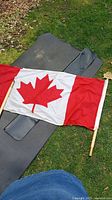 Full view of Canadian flag with wooden pole on black carrying case lying on grass outdoors.