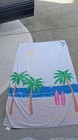 Full view of Kate Spade beach towel laid out, showing design of palm trees, ocean, sun, and surfboards.