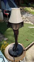 Full view of leg lamp showing black boot base, white fabric lampshade with black fringe, power cord, and outdoor backdrop.
