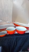 Photo showing three enamel cast iron cookware pieces: two round pots with white interiors and lids, and one shallow round dish, all flame orange red.
