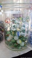 Clear glass jar filled with many colored marbles of varying sizes including green, clear, white, and orange marbles. Some plastic bags with marbles visible beside the jar.