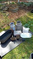 Full view of lot outdoors on black mat showing all 8 items including 3 milk cans, 2 brass pots, 3 metal planters in various finishes with rust and wear.