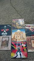 Seven vinyl record albums arranged outdoors on concrete, visible Iron Maiden, Judas Priest, Black Sabbath, Led Zeppelin covers.