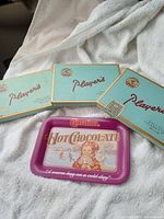 Three vintage Player's Navy Cut Mild cigarette tins and one Carnation Hot Chocolate metal serving tray on white fabric surface.
