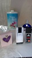 Four fragrance items displayed on white surface: Vera Wang Princess perfume in pink box, Bling by Mariah Carey perfume, Adidas Team Force body spray, and Freesia Talc powder container.