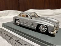 Side view of silver Mercedes 300SL display model car on gray base with ruler for size reference.